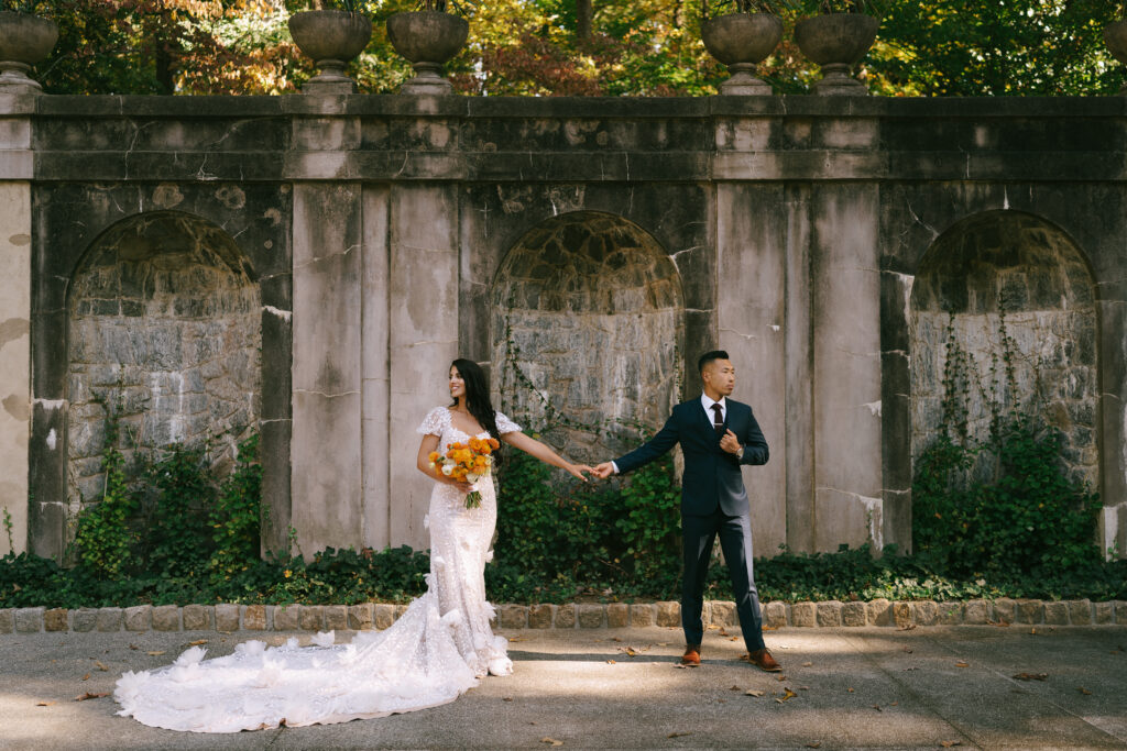 Bride and Groom stand with fingers touching, looking away from one another and facing the viewer. A large stone wall covered in green ivy frames the background.