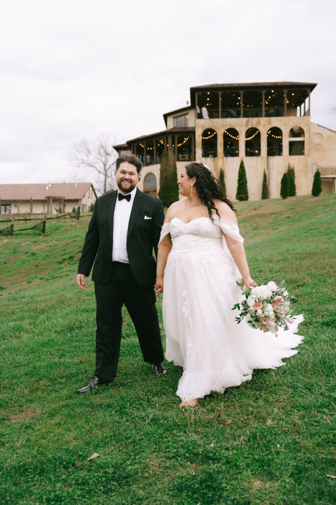 Bride and Groom walk hand in hand on green grass with a beige, multi-tone, Tuscan style, three story building in the background.