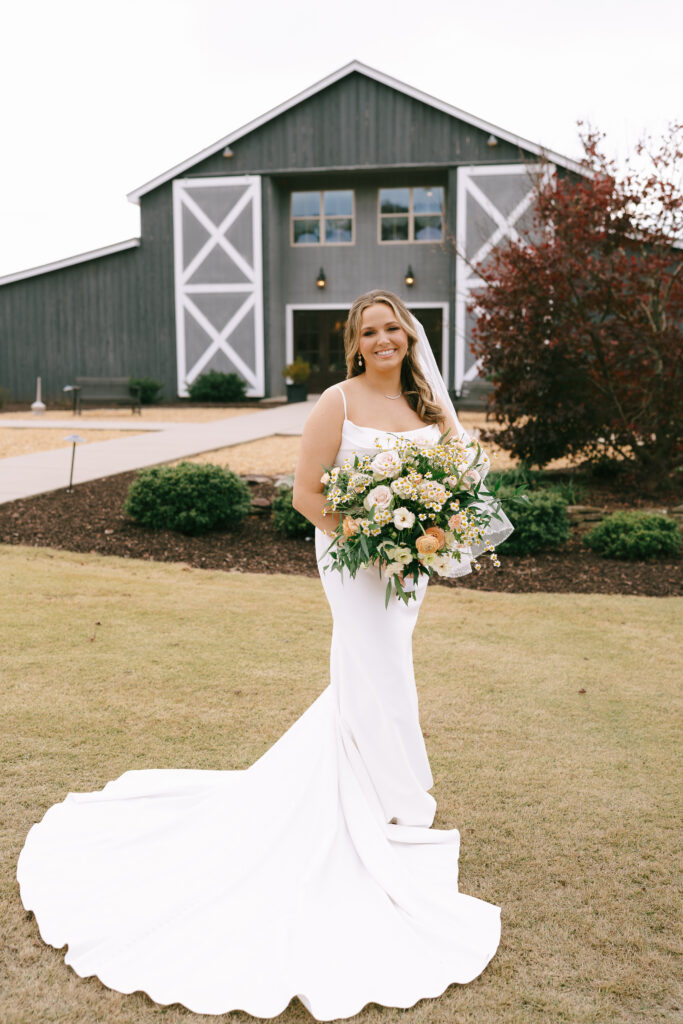 Bride holding a bouquet of colorful flowers gazing at the viewer with her dress's train fanned out in front of her and a large, grey barn in the background.