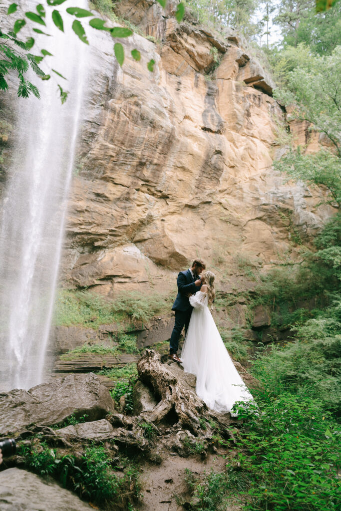 Bride and groom, standing, kiss atop large boulders. A waterfall cascades to the left and a rock face frames the background.