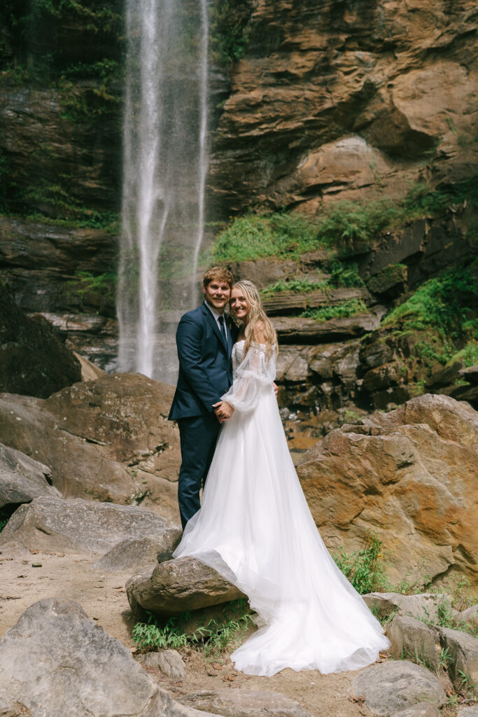 Bride in a flowy white gown and groom in a navy blue suit stand hand in hand on a large boulder facing the viewer. A waterfall cascades in the background.