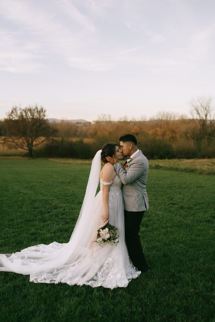 Bride and groom kiss in an open field with green grass and a setting sun creating a golden background.