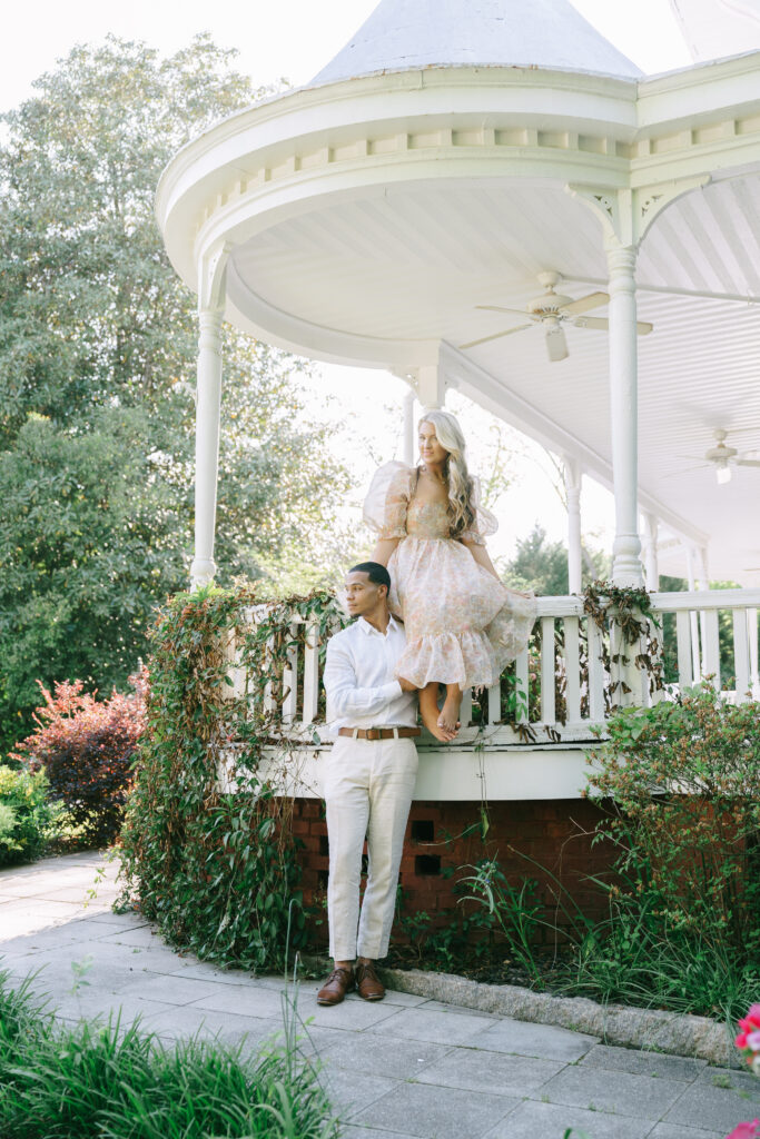 Woman in a long sleeve cream colored dress sits on the fencing of a rounded patio with a cone shaped roof. Ivy crawls up the fencing beside a man in a white collared shirt with khaki pants who stands to the left of the woman and holds her ankle.