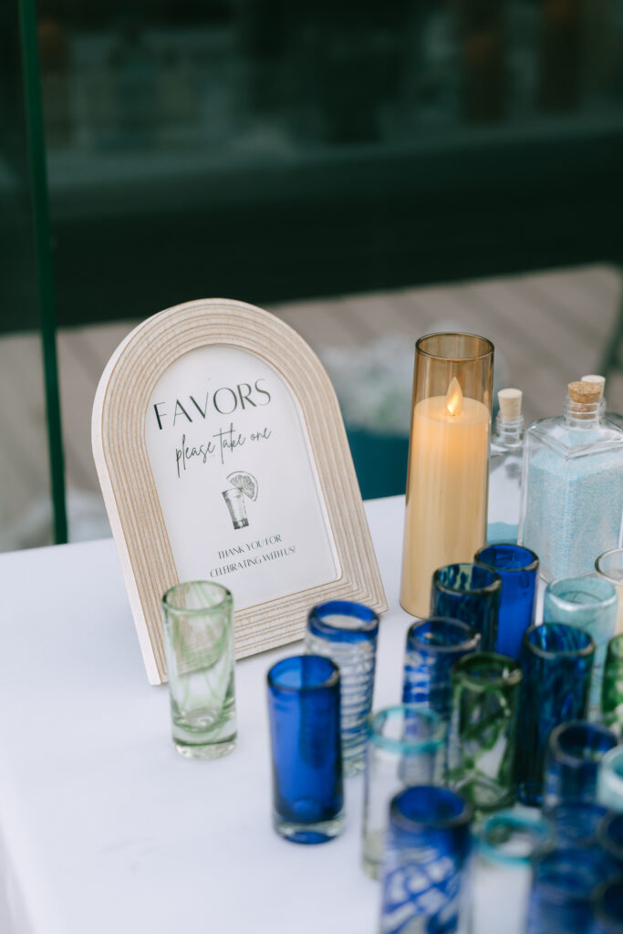Close up of arched white-washed wooden sign that says Favors with a flameless candle in a peach colored glass to the right. Blue, green, and clear shot glasses in various patterns sit in the foreground.
