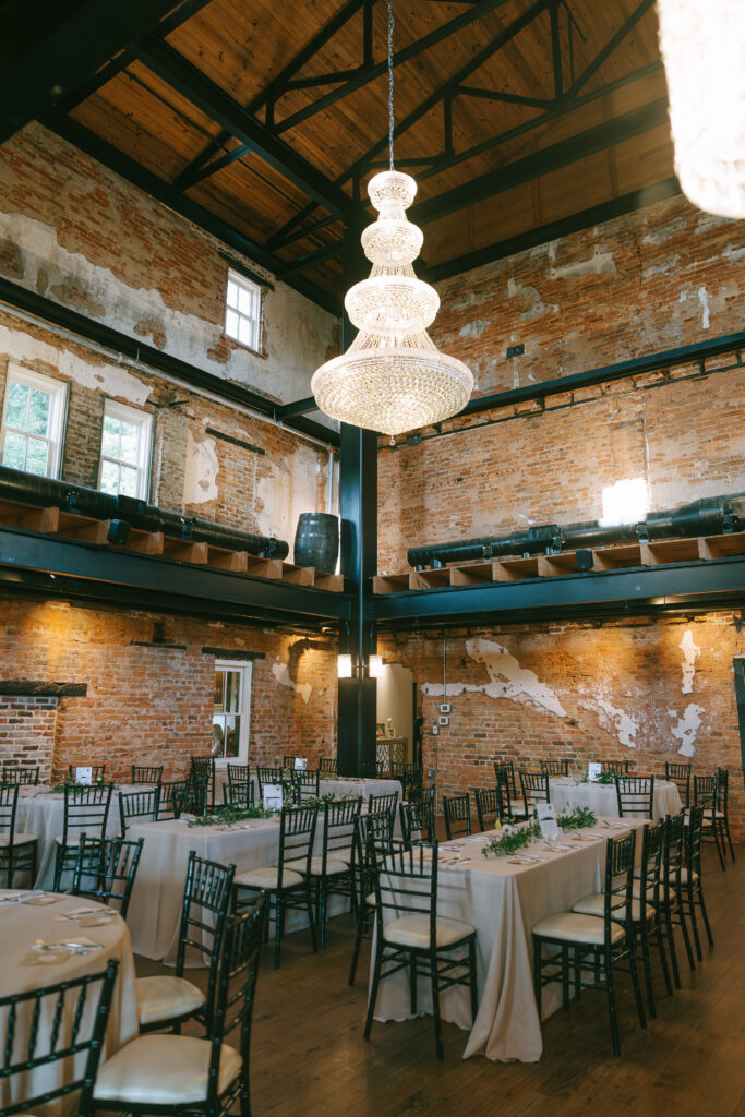 A large four tier chandelier hangs in the middle of a brick room supported by metal beams. Rectangular tables with black chairs sit on a concrete floor. 