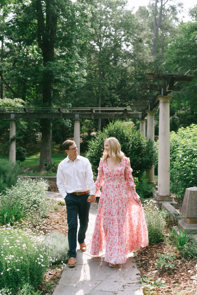 Man in a white collared shirt, blue pants, and brown shoes gazes at a woman in a long sleeve, pink, floral dress on a stone path surrounded by lush green trees. 