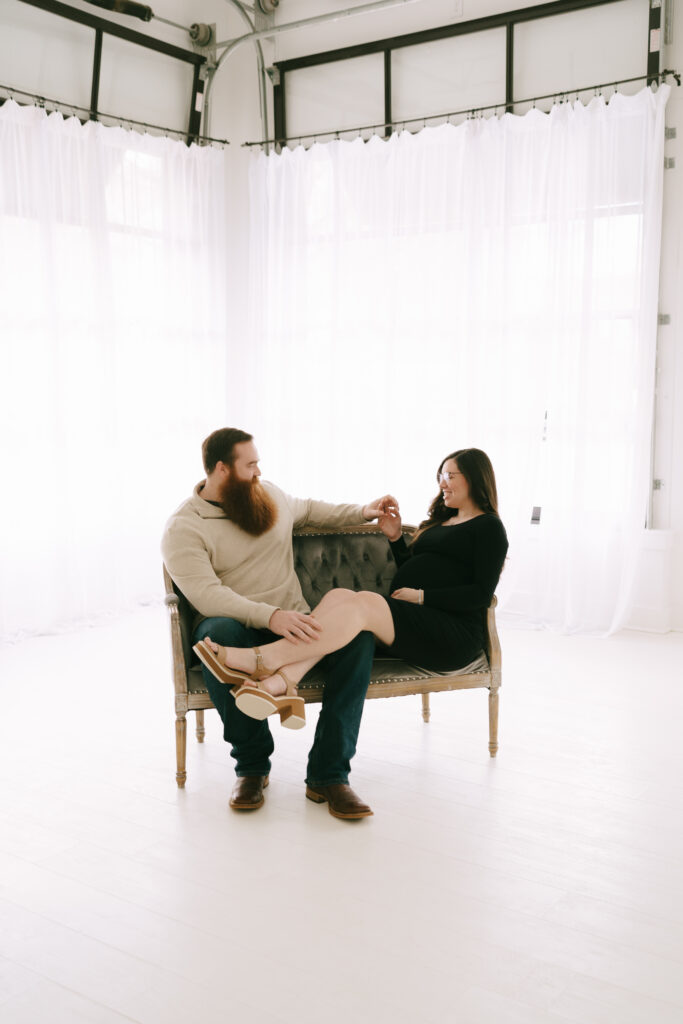 Couple lounging and gazing at one another on a small loveseat in a whitewashed room