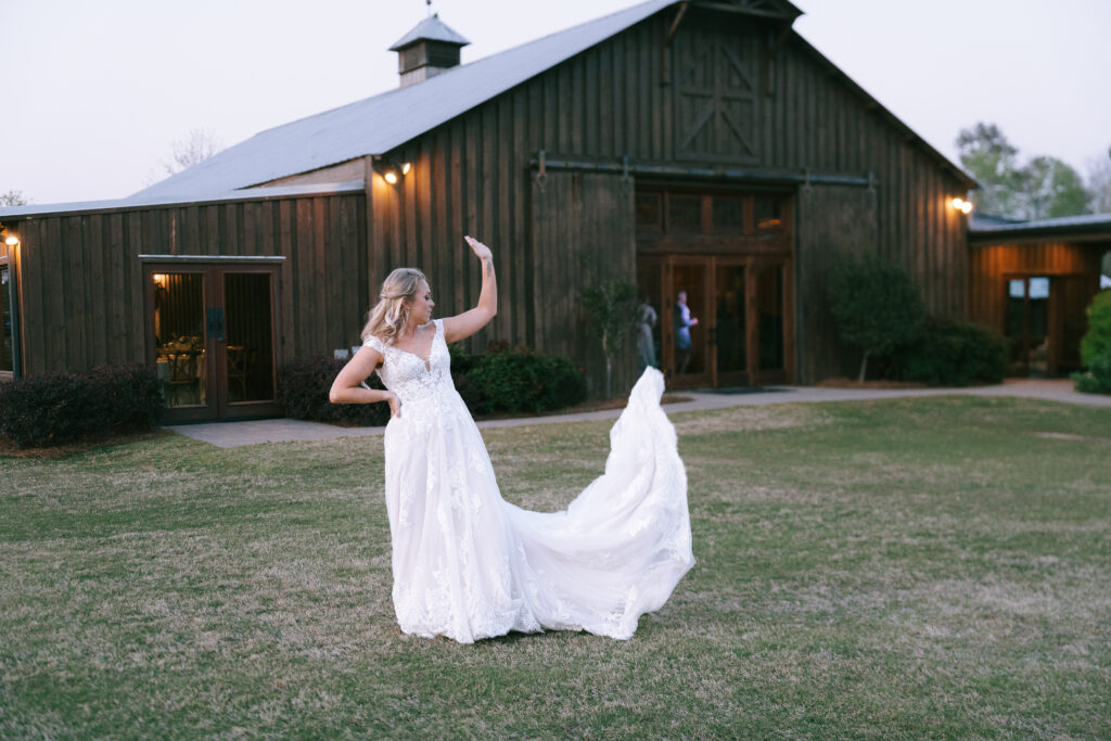 Bride flicking her wedding dress train with a hand on her hip in front of a wooden barn with rolling doors.