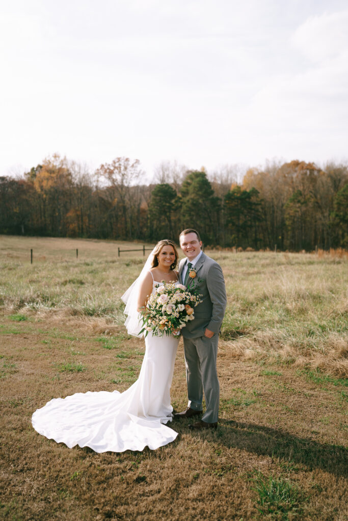 Bride and Groom standing in a field with golden grass and yellowing fall trees in the background.