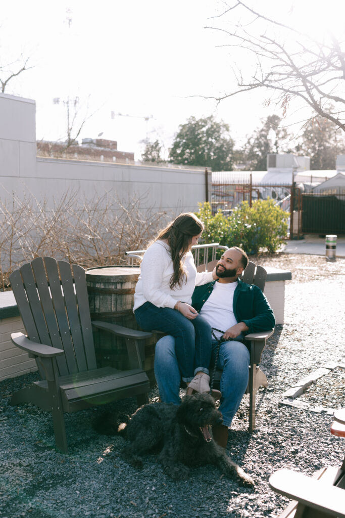 Couple sitting together in an Adirondack chair on a gravel outdoor space with their black dog