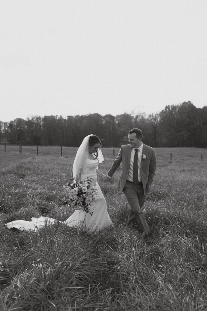 Black and white photo of bride and groom walking through a field, hand in hand.