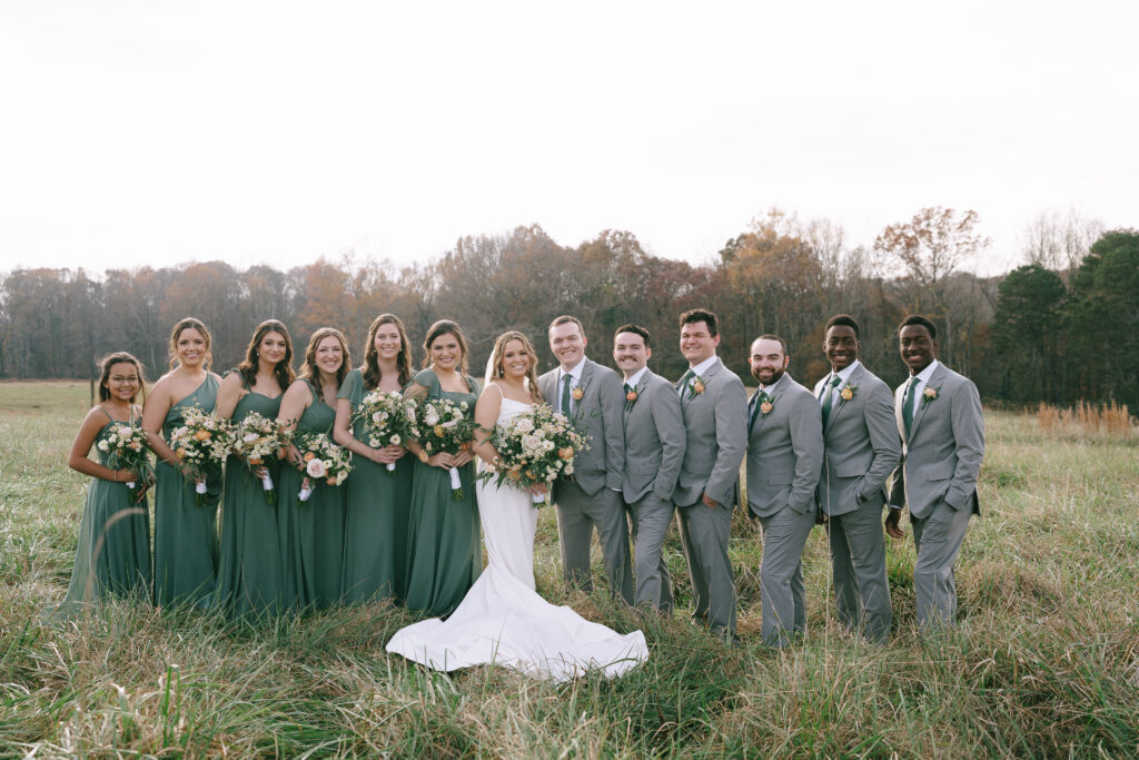 Bridesmaids in green dresses holding bouquets of flowers to the left, bride and groom in the center, and groomsmen in grey suits with green ties to the right all standing in a golden grass field.