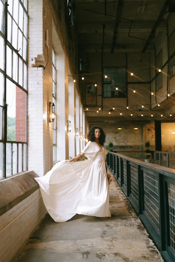 Bride waves her dress on a concrete and brick balcony with metal railing, exposed metal beams and bistro lights overhead.