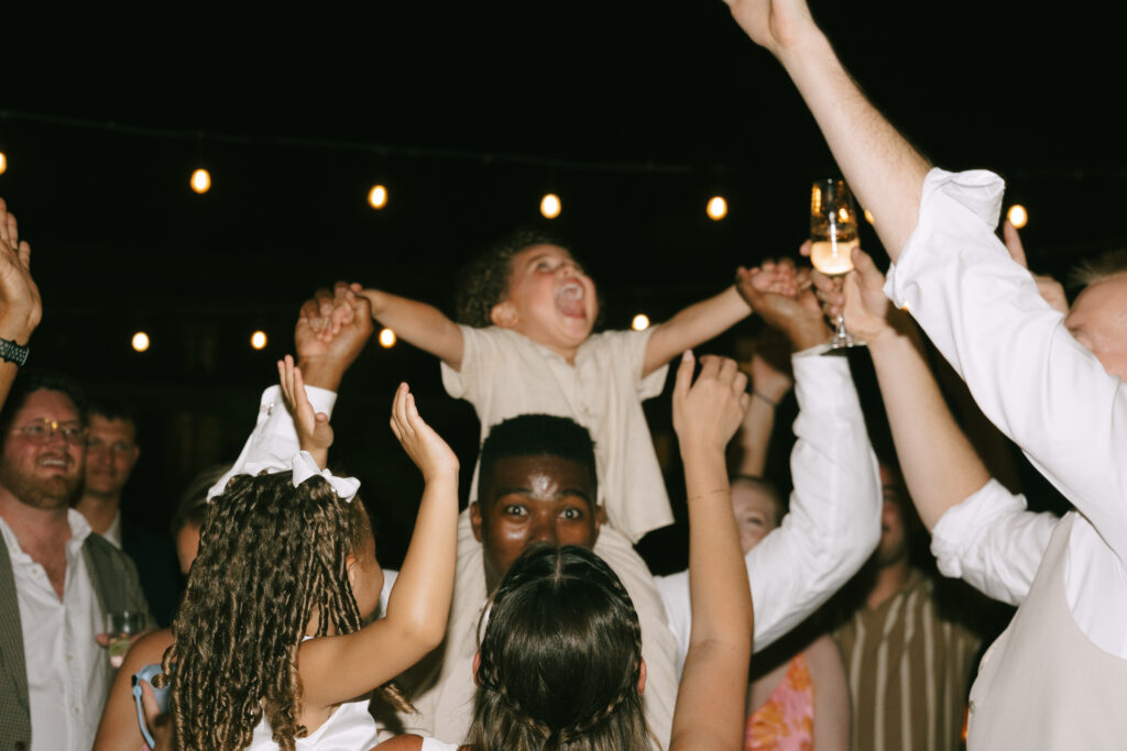 Group of people dancing with a close up of child sitting on shoulders with arms spread laughing to the sky.