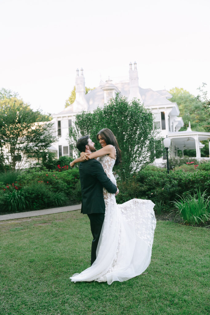 Groom lifts bride by her waist and Bride gazes at Groom with arms wrapped around his shoulders. They stand on a green lawn with green bushes and trees surrounding them. A white manor house sits in the background.