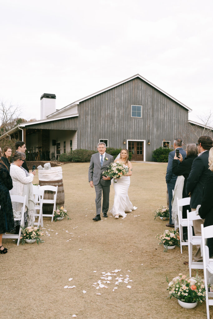 Bride and Father walk down a wedding aisle with a grey farmhouse in the background.