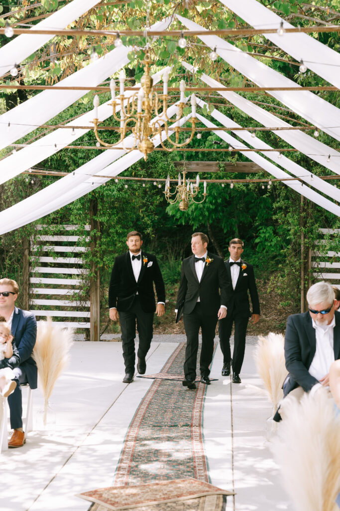 Groom and groomsmen walk down a carpet covered aisle with white drapery and gold chandeliers overhead, and deep green trees cover the backround.