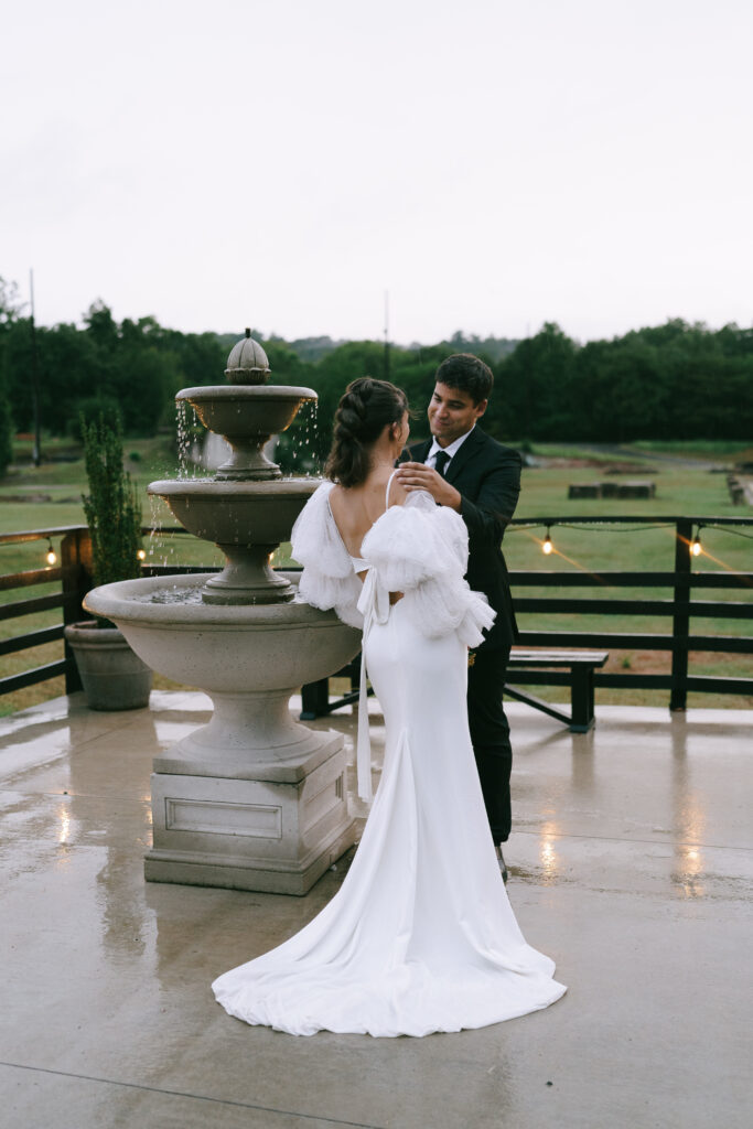 Bride and groom lean on three tiered circular fountain and gaze at each other. Bride faces away from the viewer.