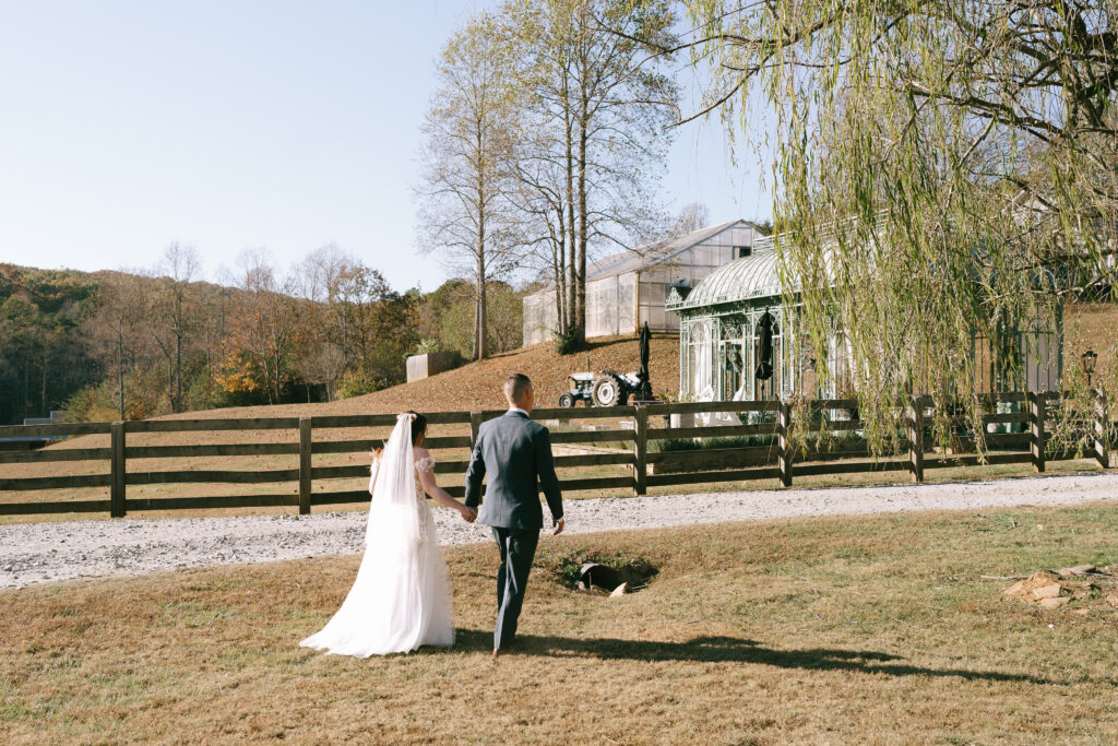 Bride and groom walk hand in hand away from the viewer towards a green, oxidized greenhouse surrounded by a wooden fence.