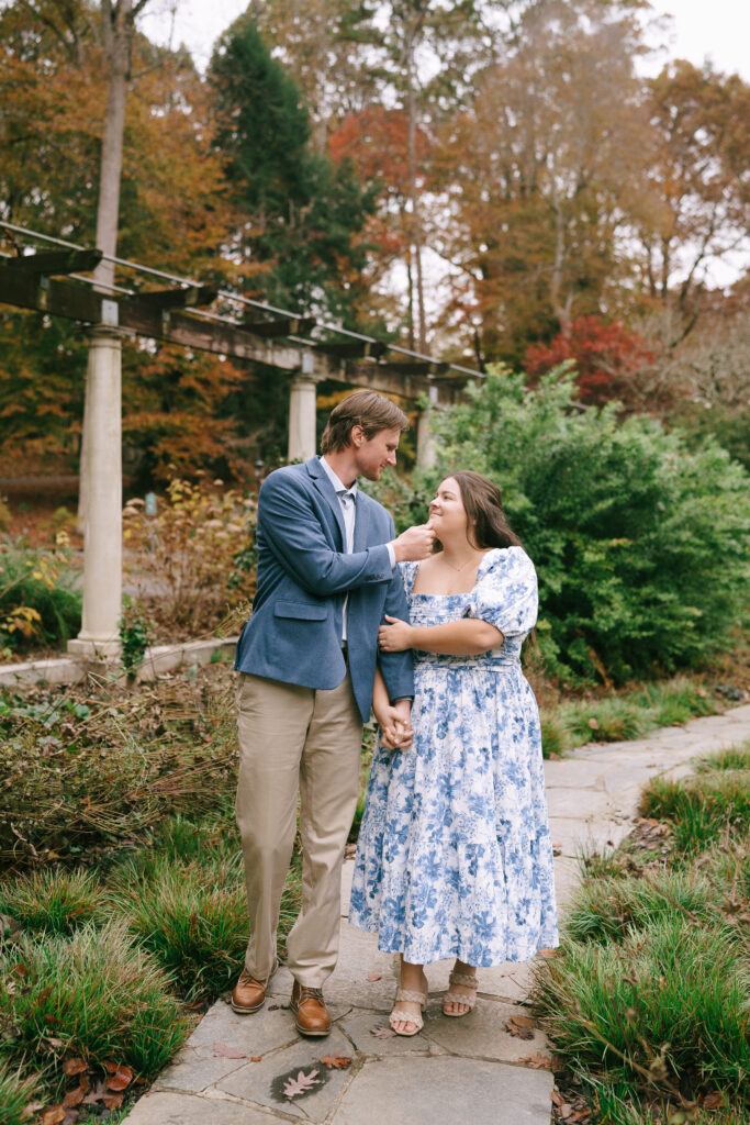 Man in a blue suit jacket, khaki pants and brown shoes cups the chin of a woman in a blue and white floral dress. They walk towards the viewer on a stone path in the middle of an autumn colored garden.