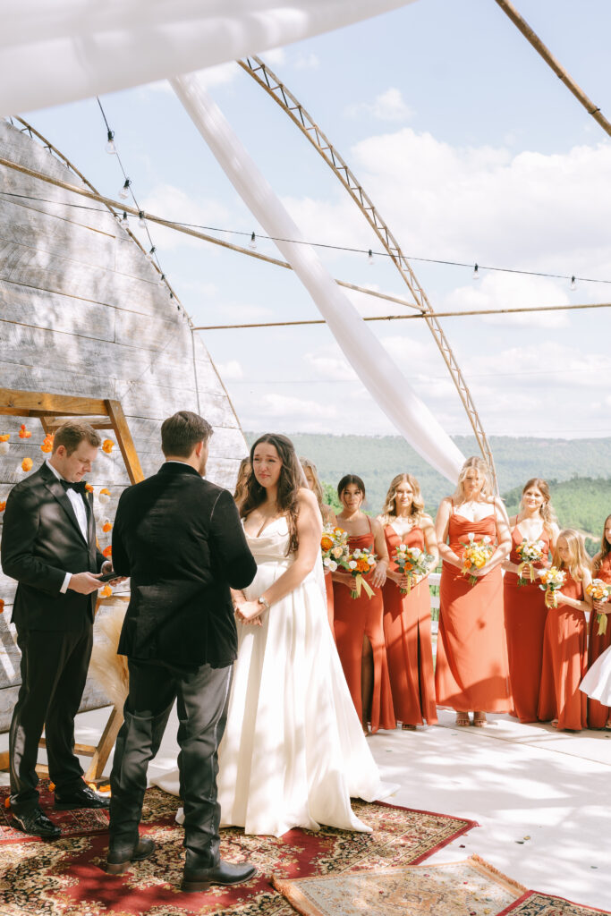 Bride and Groom stand hand in hand at the alter with bridesmaids in terracotta dresses and colorful bouquets stand in the background. The blue sky and white, puffy clouds contrast with the green mountains in the distance.