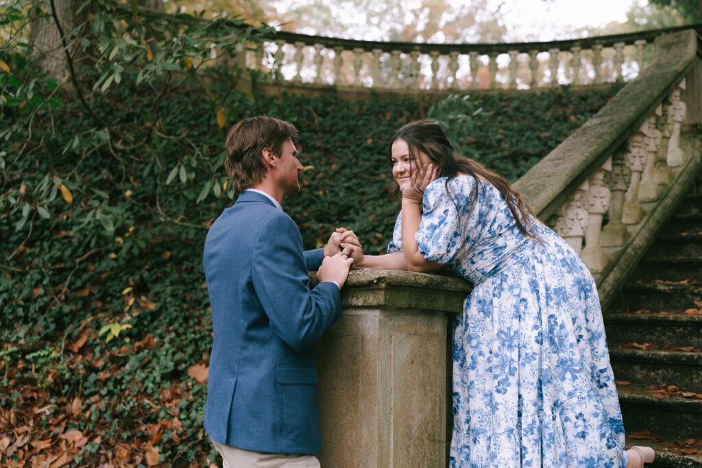 Woman in a blue and white floral dress stands with fingers intertwined with man in a blue suit jacket on a stone, ivy covered stairway.