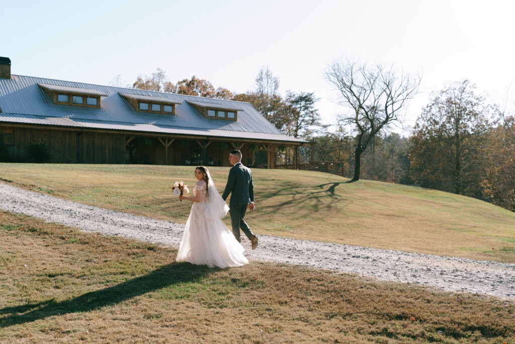 Bride and Groom walk hand in hand away from the viewer on a gravel path with a rectangular wooden building with a metal roof in the background.