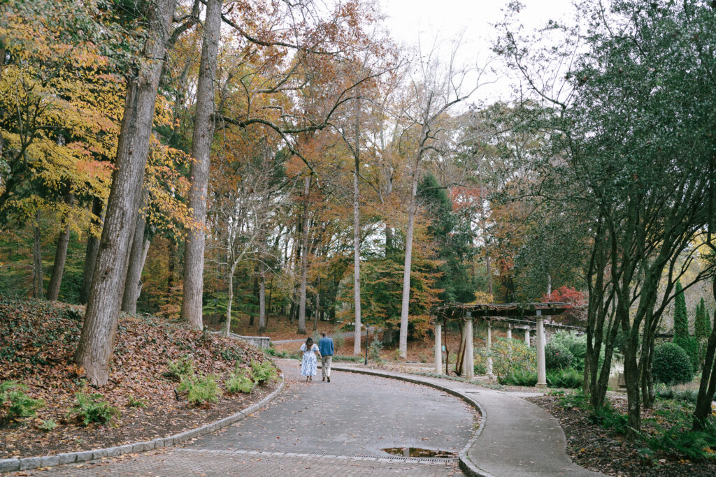 Man in a blue suit jacket and woman in a blue and white floral dress walk far from the viewer on a cobblestone road towards autumnal trees in the background.