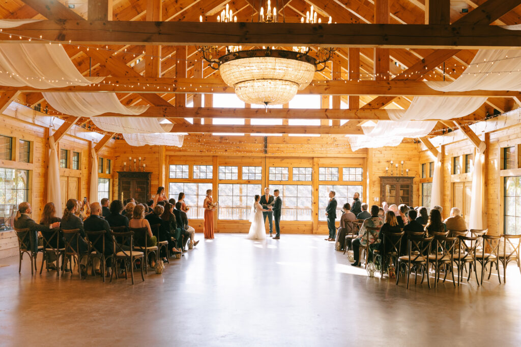 Bride and groom stand at the alter with family and friends to either side in a golden, wood toned room with white drapery between the ceiling rafters. 