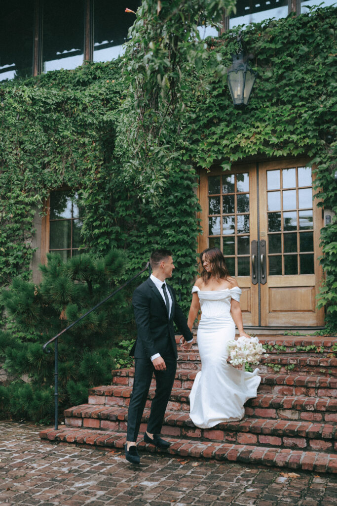 Groom leads bride down a brick staircase with ivy covered walls in the background.