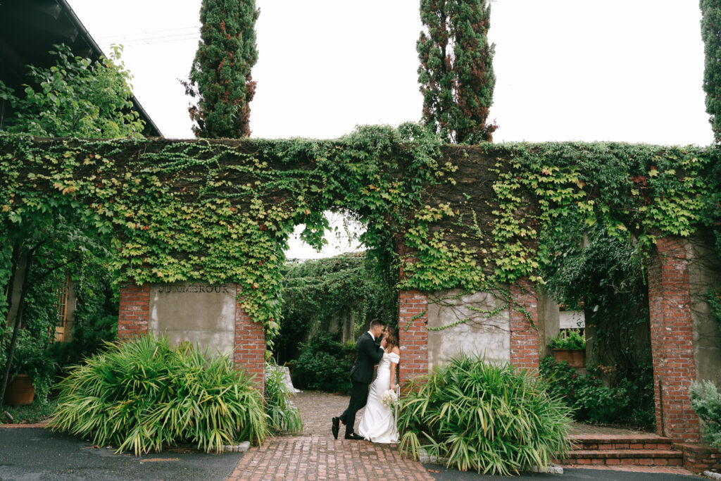 Groom leans in to kiss bride underneath an ivy and brick archway.