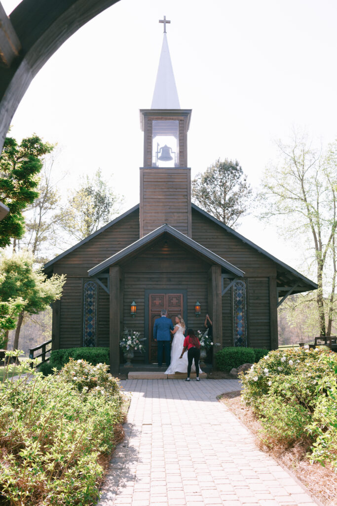 A triangular wooden church with a large steeple and bell in the background with a greenery filled walkway.