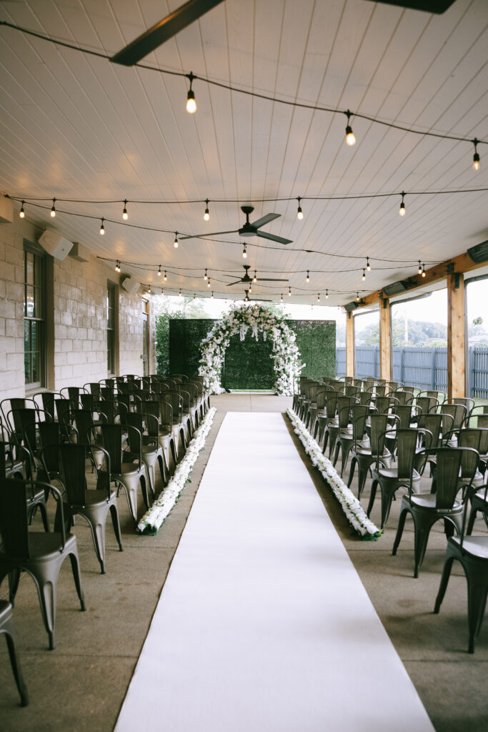 A wedding aisle with a white runner leads to a white floral arch. White wooden paneling and bistro lights above.