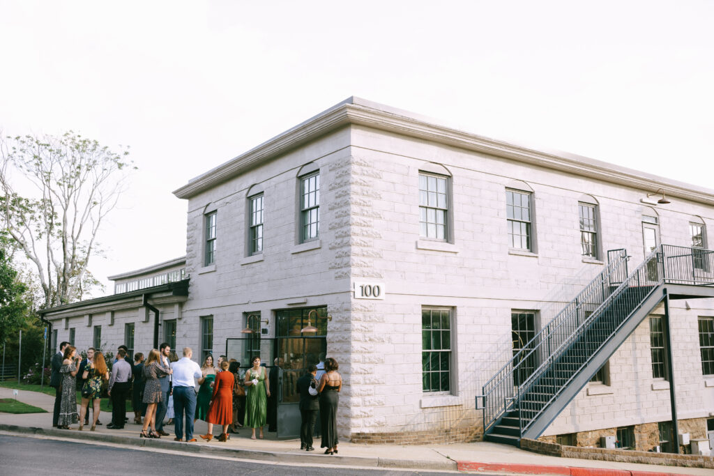 White stone building with the number 100 and a metal staircase to the right. Men and women gather at street level outside.