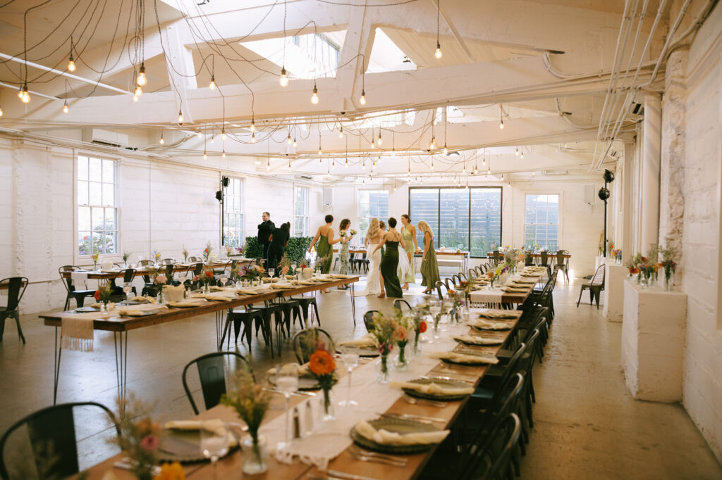 Long rectangular tables set for dinner in a white room with windows and skylights. A group of women and the bride gather in the background.