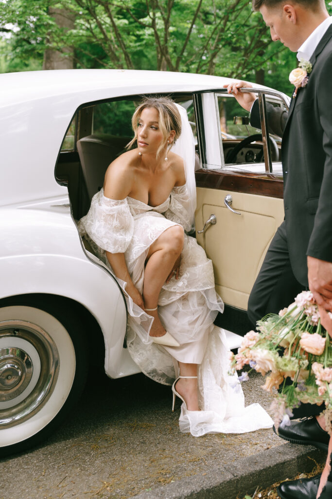 Bride fixing shoe sitting in a white Rolls-Royce, gazing into the distance