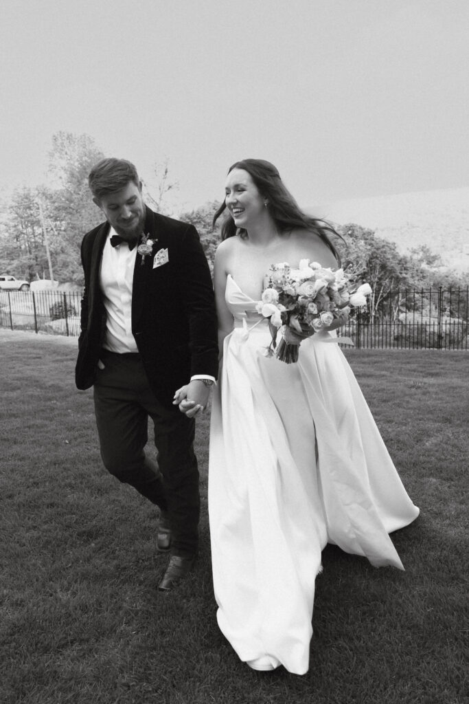 In black and white of bride and groom holding hands, smiling, and walking together.