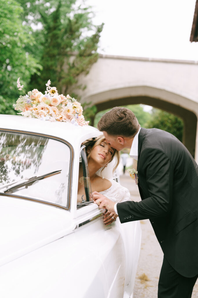 Groom kisses bride, who is sitting in a white Rolls-Royce, on the forehead