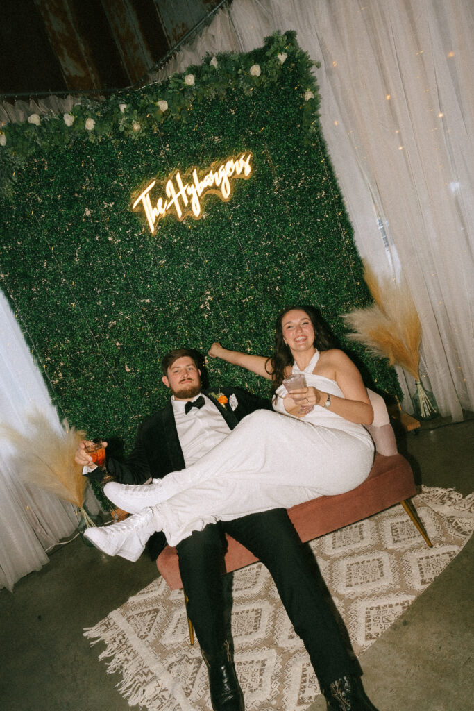 Bride and groom sitting on an orange couch in front of a faux greenery covered wall.