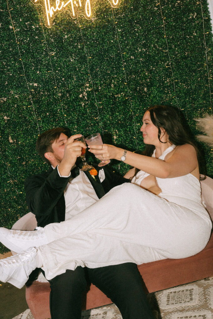 Bride and groom sitting on an orange couch in front of a faux greenery covered wall doing "cheers" with drinks.