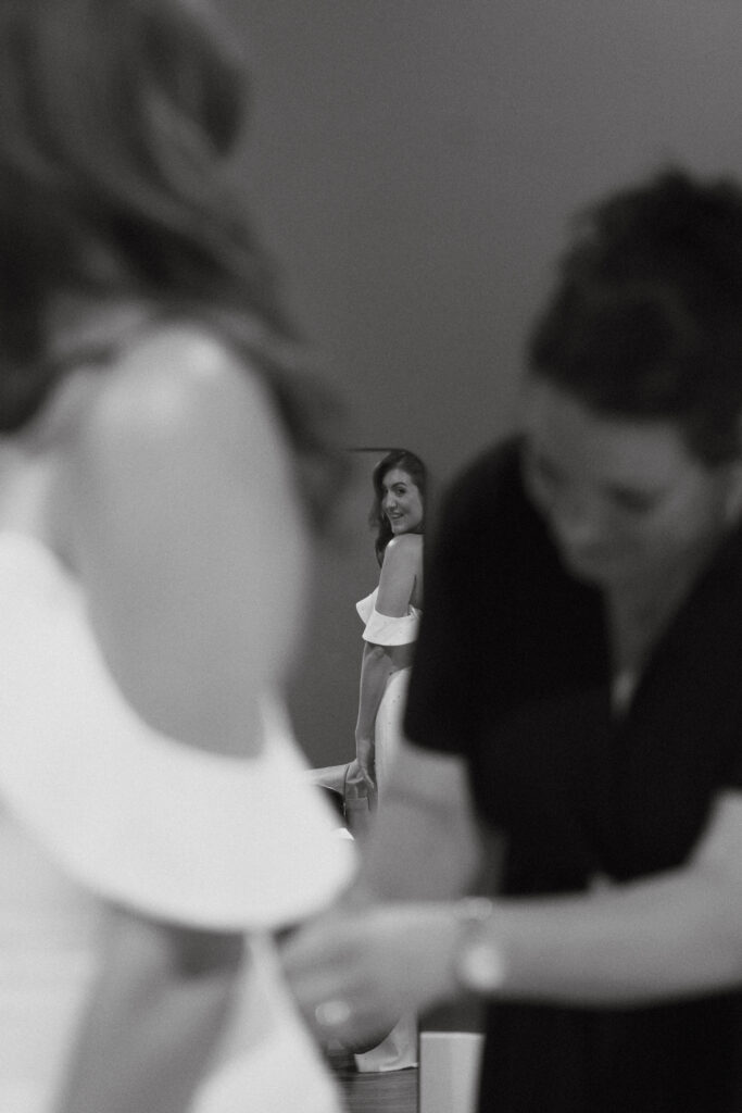 In black and white of bride staring over shoulder into mirror while mother of the bride buttons gown.