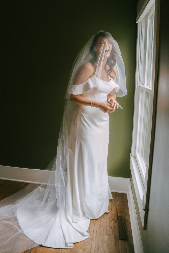 Bride in bridal gown and veil smiling and laughing in a green colored room.