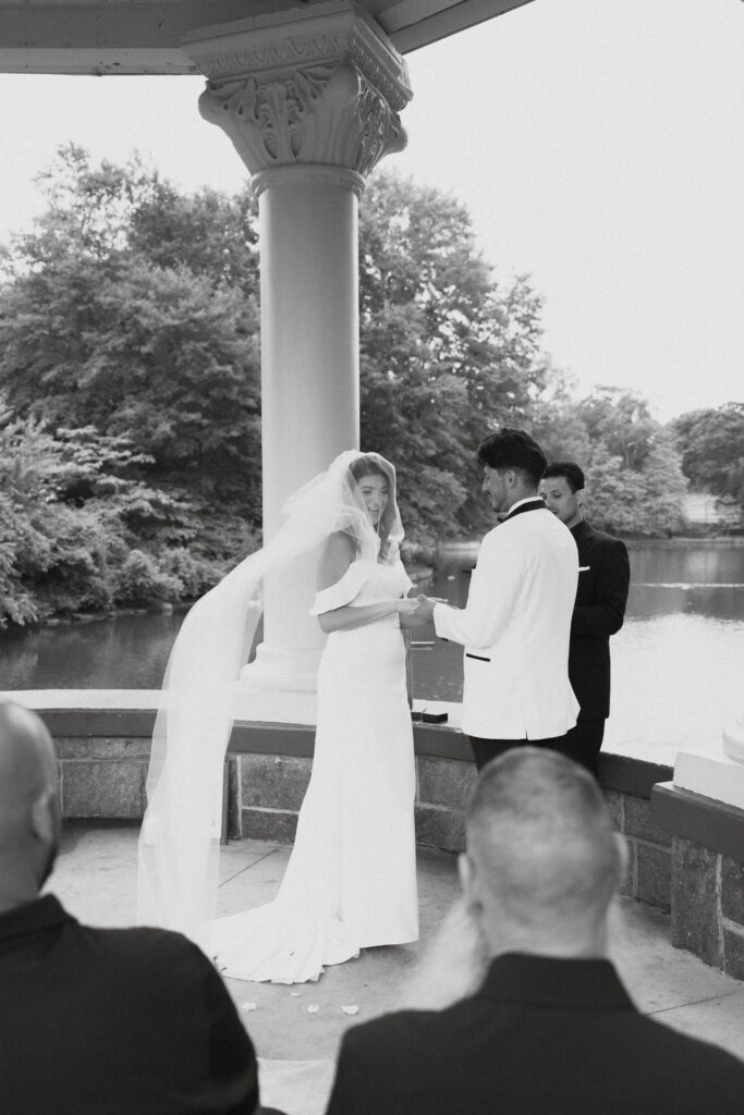 In black and white of bride and groom clasping hands at the alter while veil blows in the wind.