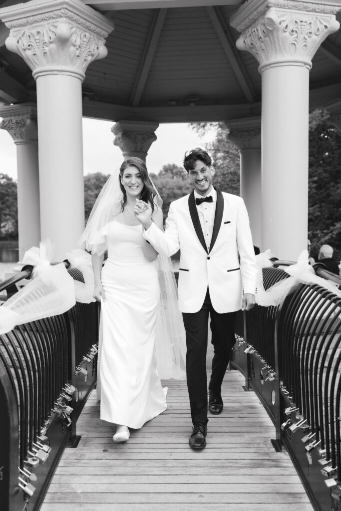 In black and white of bride and groom walking hand in hand down a lock filled bridge.