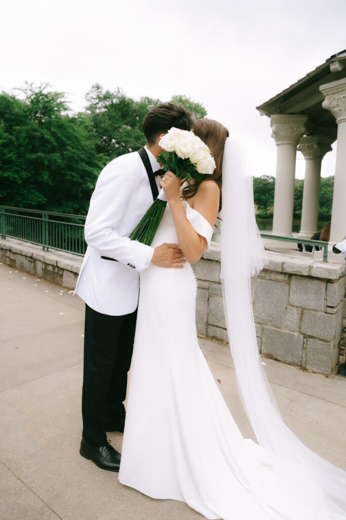 Bride and groom kissing while bride hides faces with white roses.