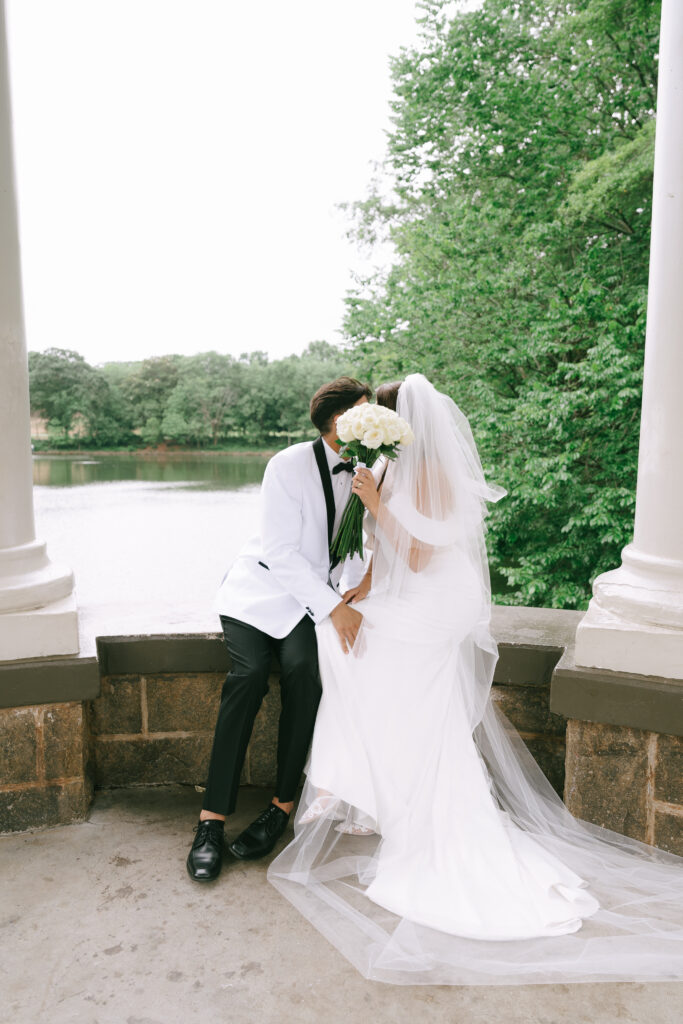 Bride and groom seated, kissing under the pavilion while bride covers faces with white roses.