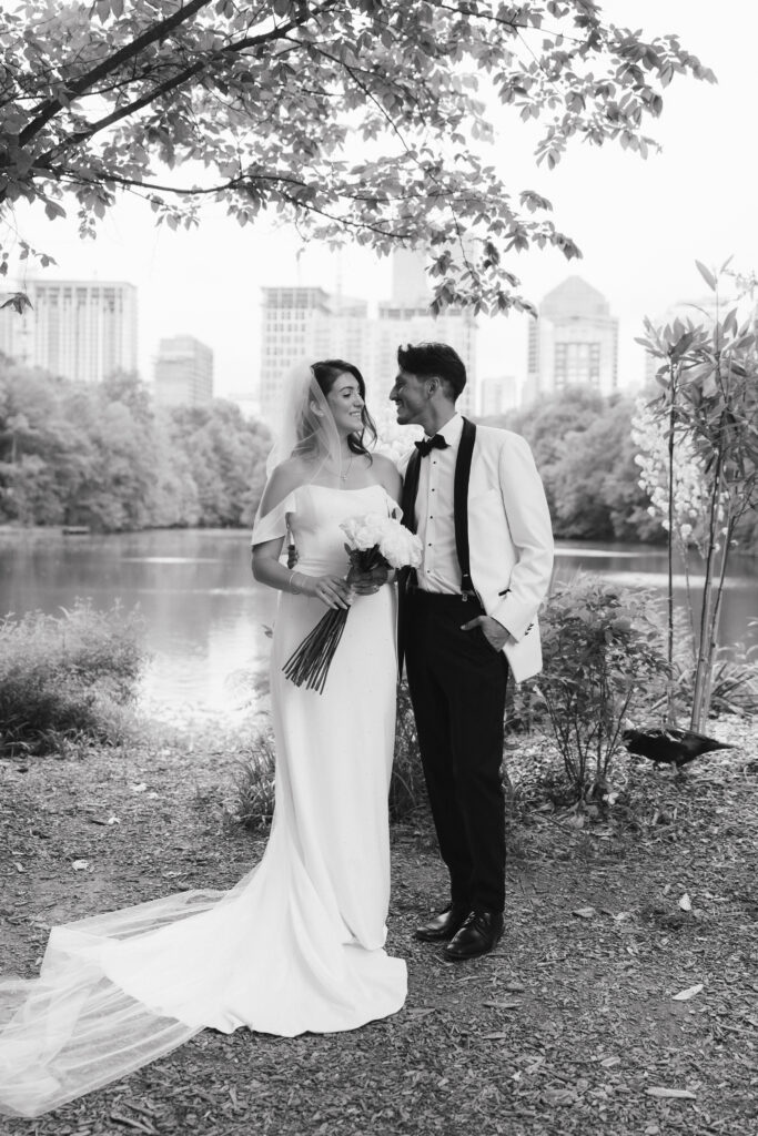 In black and white of bride and groom standing in Piedmont Park with city skyline in the background.