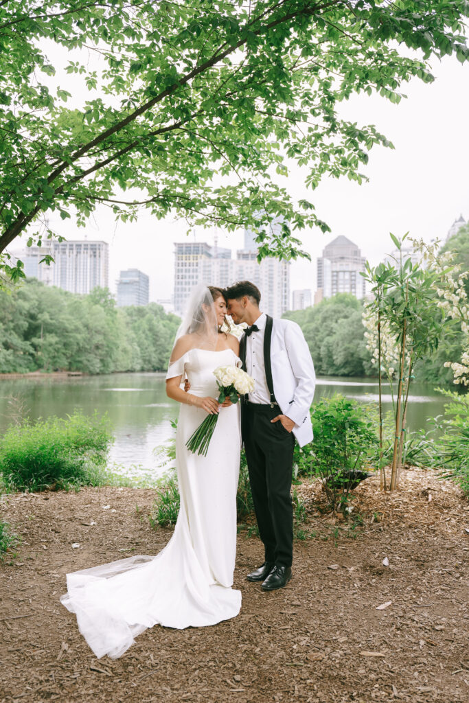 Bride and groom standing with foreheads touching in Piedmont Park with city skyline in the background.