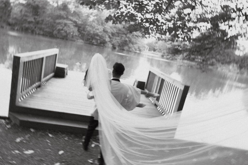 In black and white of groom carrying bride towards a wooden dock while veil blows.