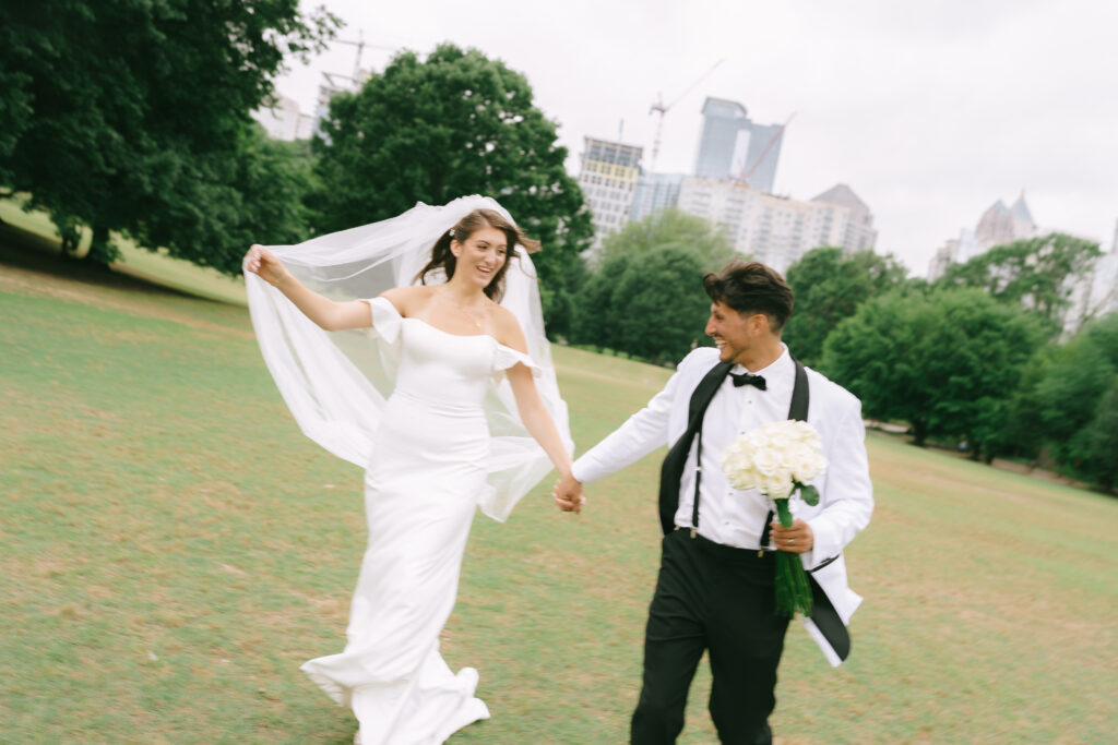 Bride and groom clasping hands and running in Piedmont Park with city skyline in the background.